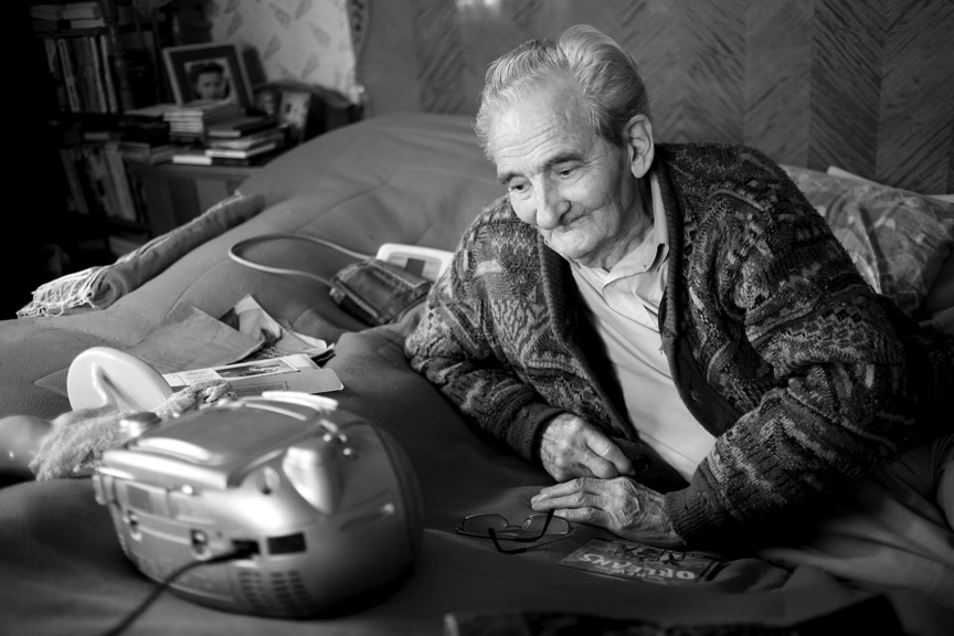 Photographie en noir et blanc avec à gauche Licu assis sur un lit. On ne voit pas son visage. Ses mains sont croisée. La main droite tient une télécommande, la gauche est posée sur le genou droit. 