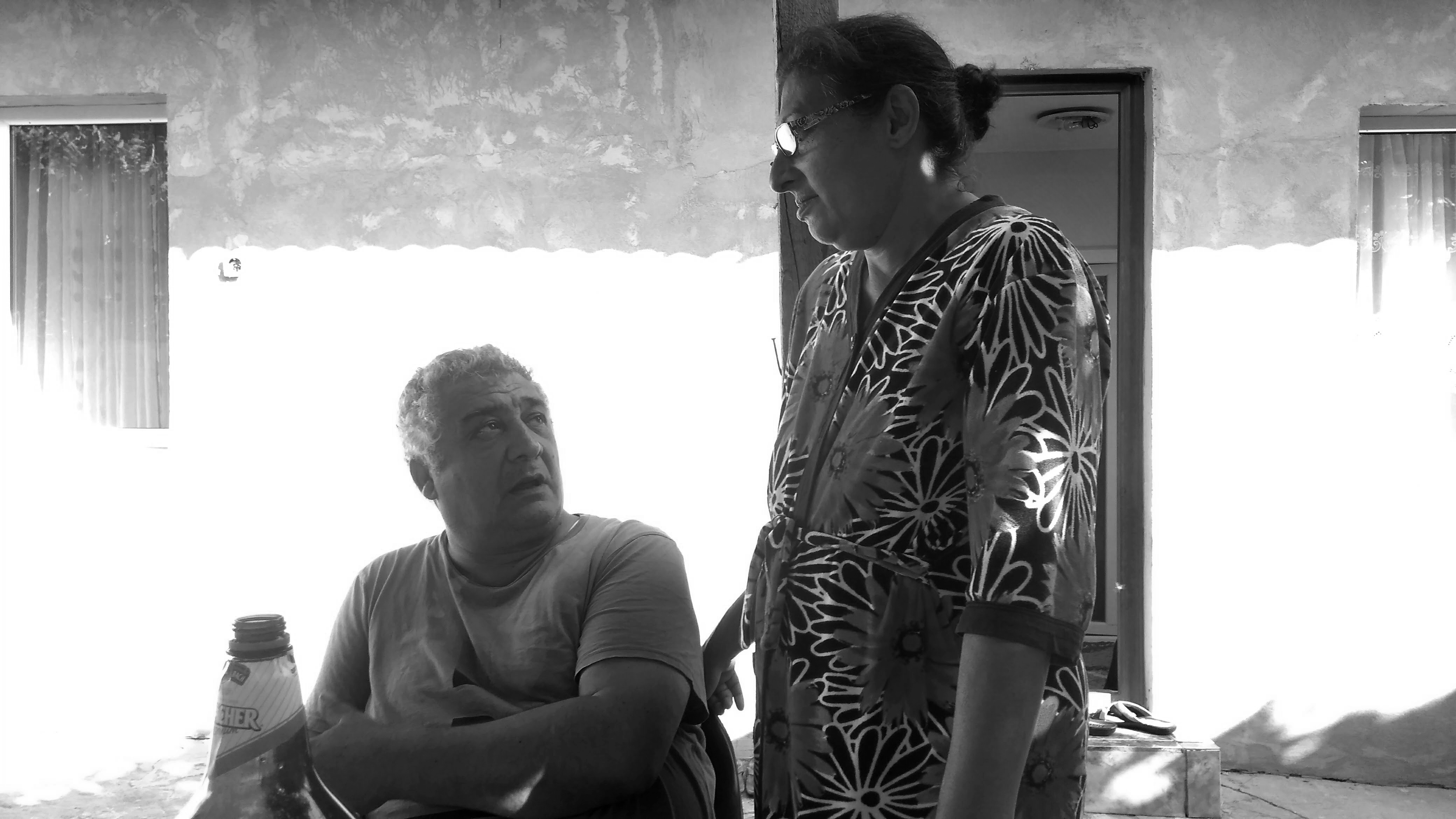 Photographie en noir et blanc de Gheorghe et Sorina, sa femme. Gheroghe est assis à une table dans la cour de sa maison et parle en la regardant. Sorina est débout, la main appuyée sur le dossier de la chaise, elle le regarde.
