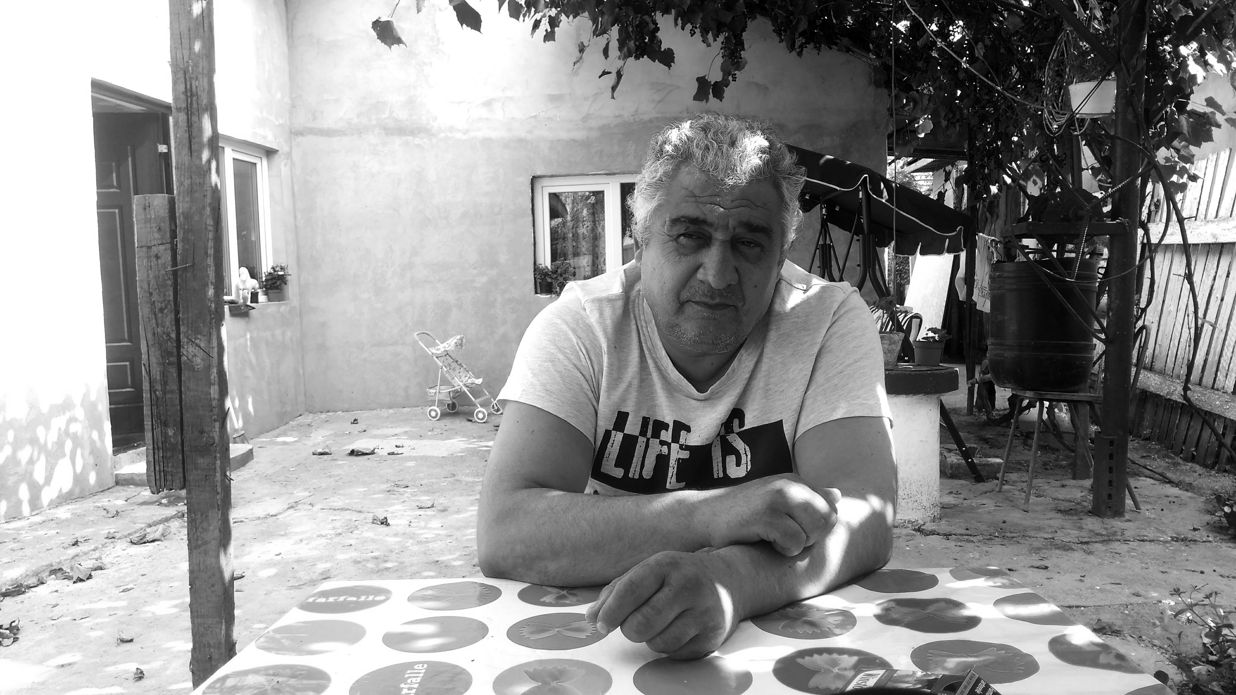 Photographie en noir et blanc de face de Gheorghe qui regarde l’objectif. Il est assis à table, dans la cour de sa maison. En arrière plan, on voit la maison et une balancelle. A droite de l’image et au dessus, il y a une vigne.