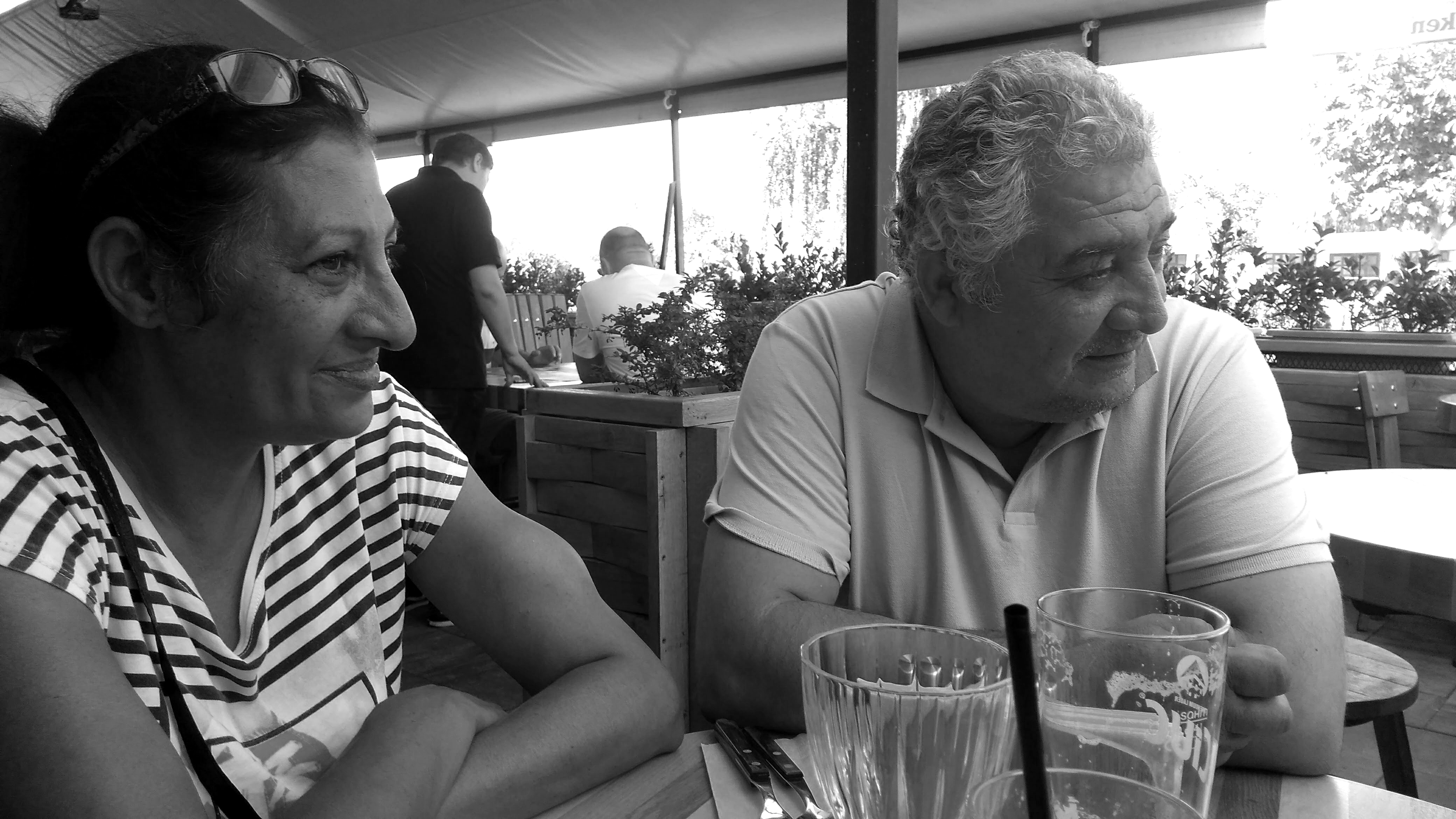 Photographie en noir et blanc de Gheorghe et Sorina, assis à la table d’un restaurant. Ils regardent tous les deux à la droite de l’image en souriant.