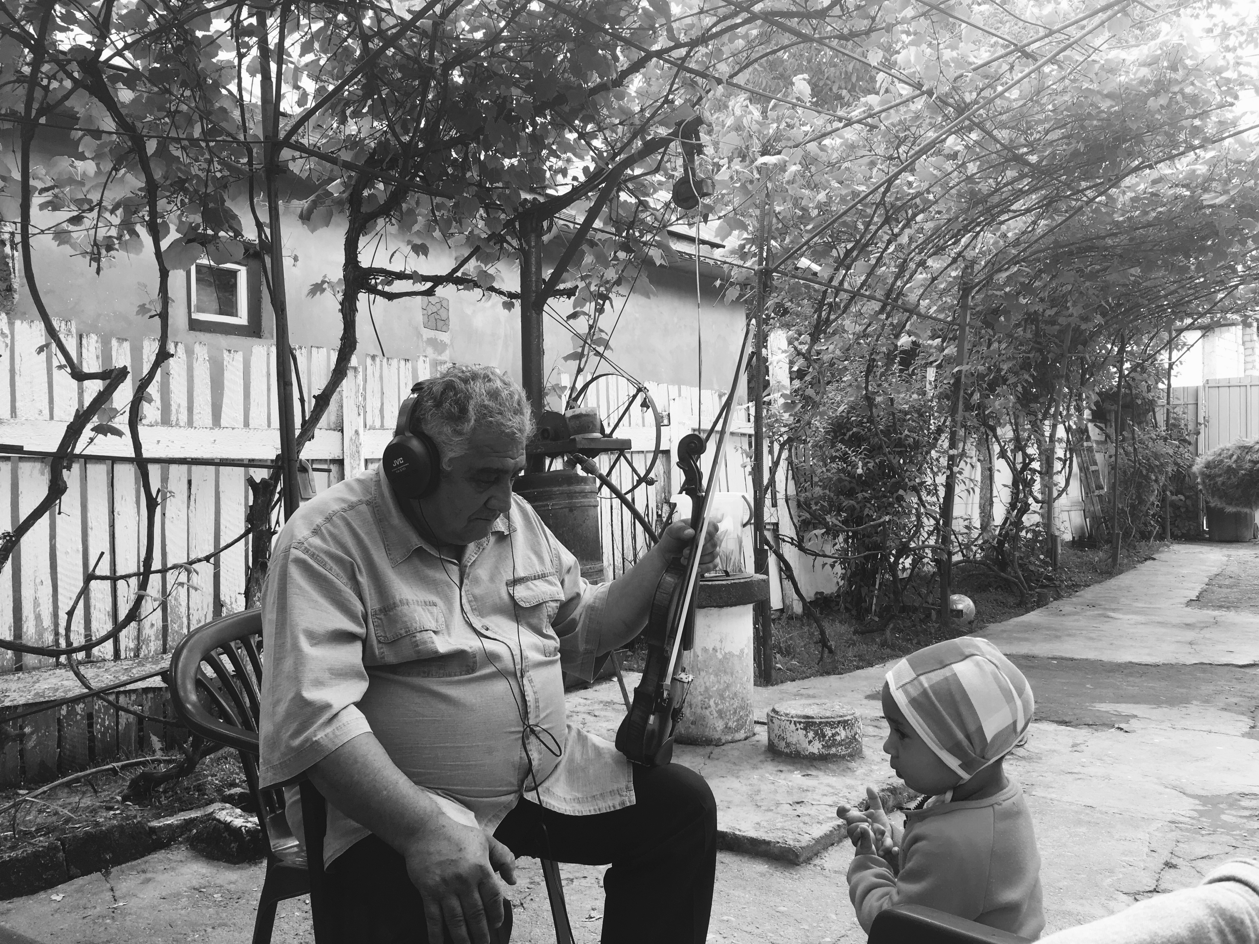 Photographie en noir et blanc de Gheorghe assis sur une chaise de jardin dans la cour de sa maison. Sa main gauche tient son violon et son archer. La droite est posée sur son genou. Il écoute attentivement à travers un casque qu’il porte sur les oreilles. En face de lui, une jeune enfant.